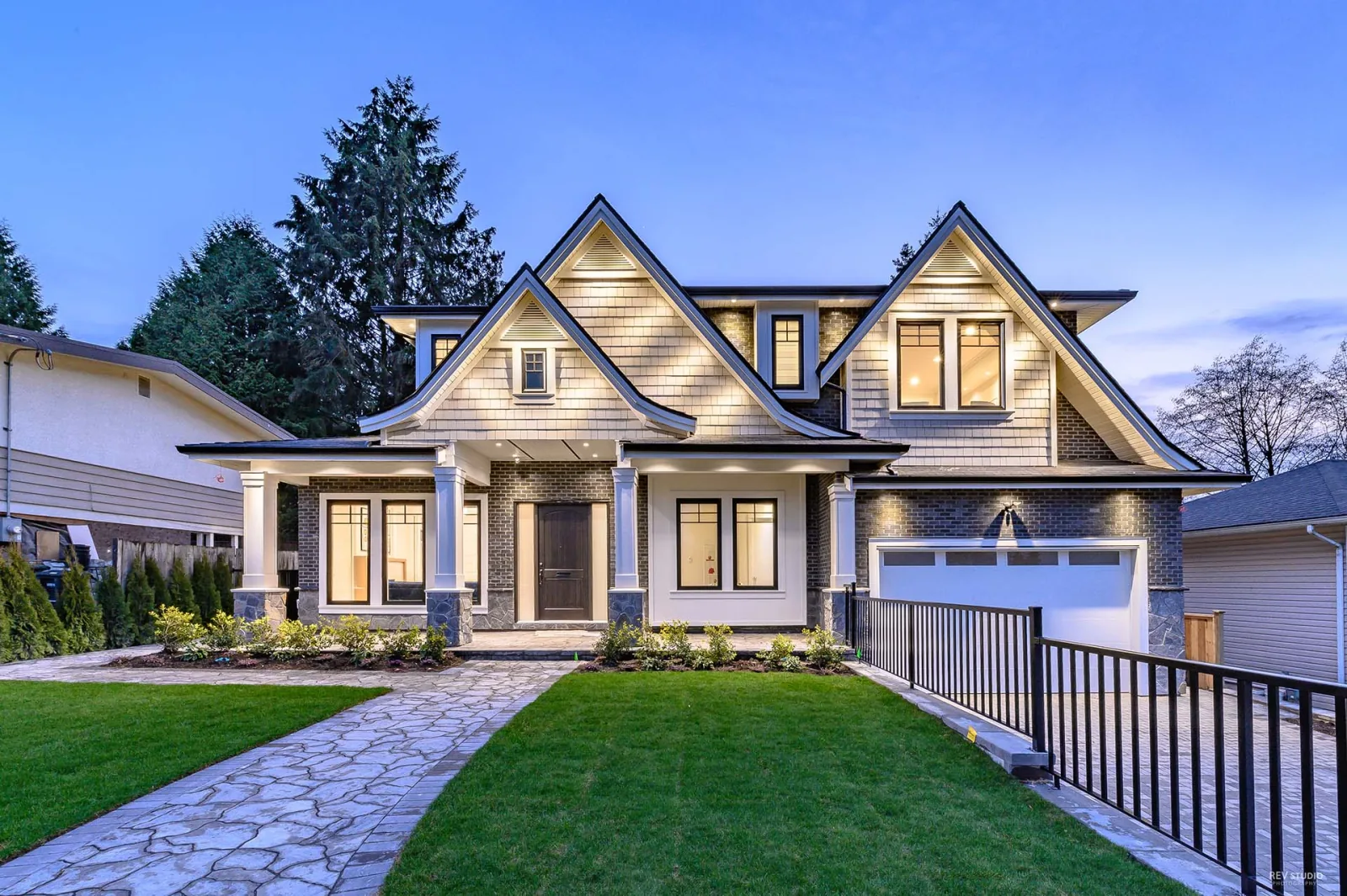 A finished custom home on 6th Street in Burnaby by Majack Developments, photographed at dusk, with three lit front gables, a stone path and a garage on the right