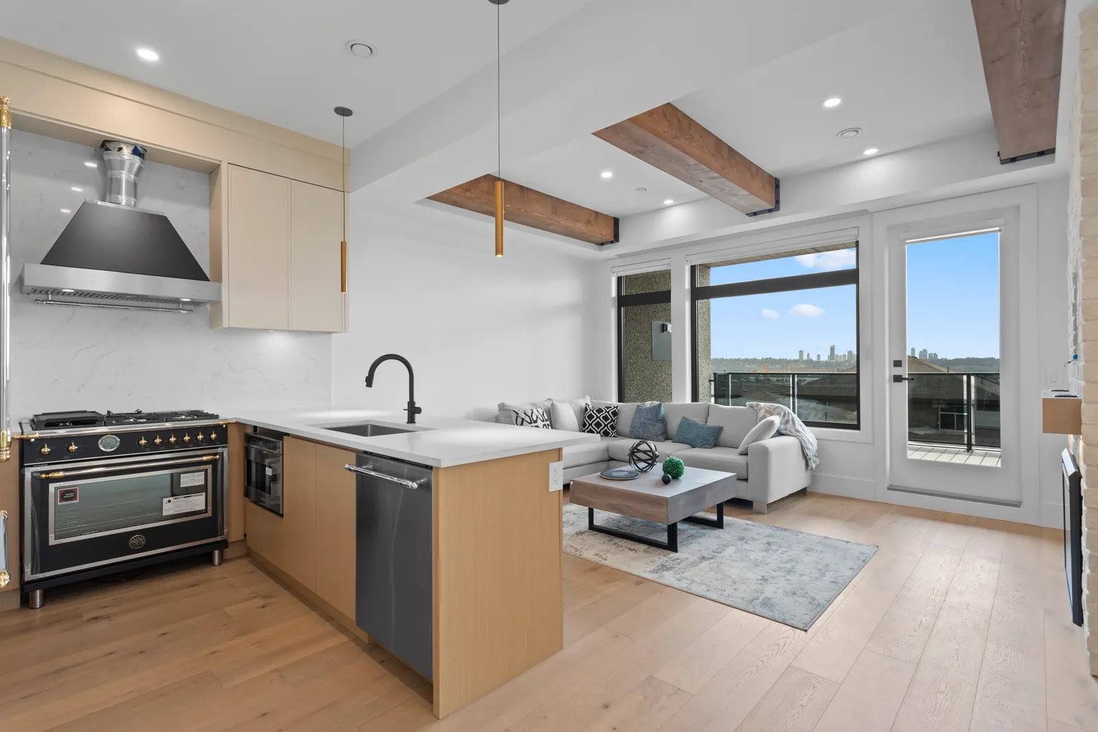The kitchen and living room of one home in the Majack Developments fourplex on Delestre Avenue in Maillardville, Coquitlam, with a black range, pale wood cabinets, exposed ceiling beams and doors opening to a deck