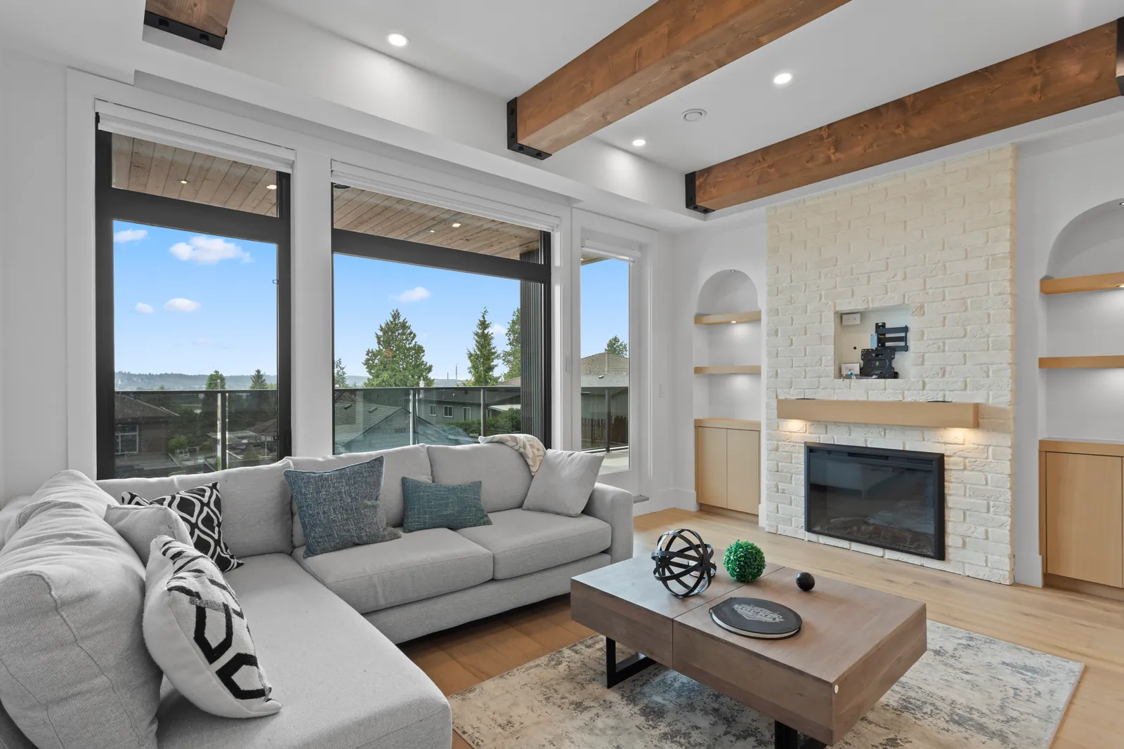 The living room of one of the four homes at the Majack Developments fourplex on Delestre Avenue in Maillardville, Coquitlam, with exposed wood beams, a white brick fireplace between two arched shelf niches, and tall windows opening to a covered deck