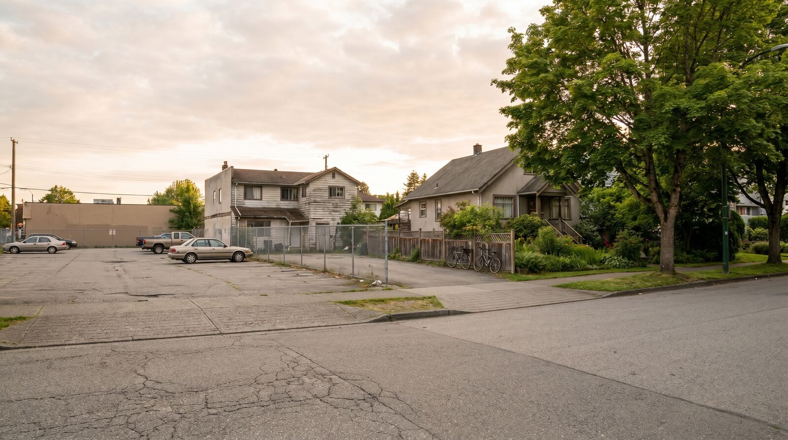 Photograph of a large surface parking lot dominating a residential lot with a small two storey building barely visible behind it, contrasted on the opposite side of a quiet Vancouver residential street with a smaller older building and street parking, late afternoon natural lighting