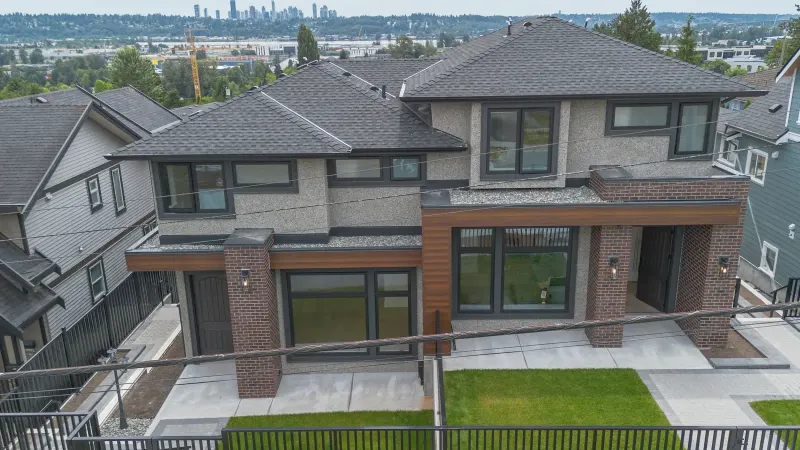 The street side of the Majack Developments fourplex on Delestre Avenue in Maillardville, Coquitlam, seen from above, with two front entries, brick columns, fenced front lawns and the Burnaby skyline in the distance